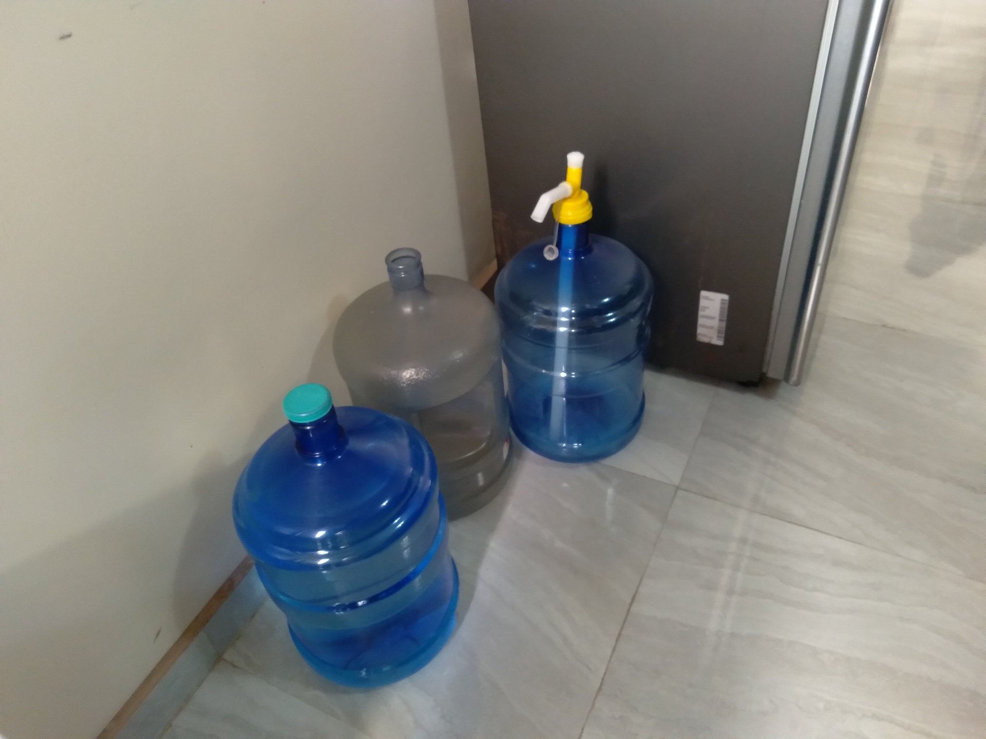 Three large water cooler bottles lined up on a tile floor against a white wall, with two blue bottles in front and one clear bottle in back.