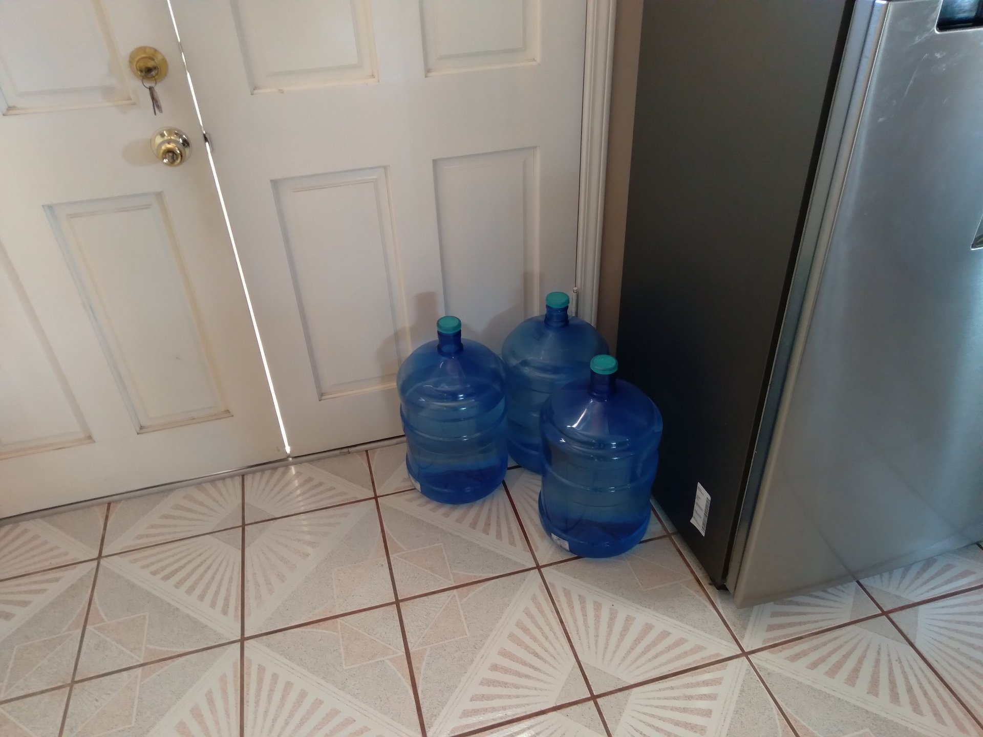 Two large blue water cooler bottles on tiled kitchen floor near white door and stainless steel refrigerator