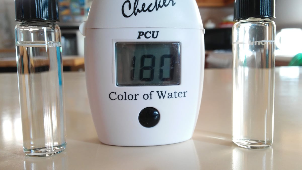 Water quality checker device displaying 180 PCU color reading, flanked by two clear glass samples on a white surface