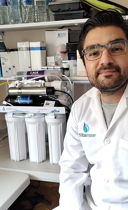 Man in white lab coat with STEMwater logo standing next to water filtration system equipment in laboratory setting