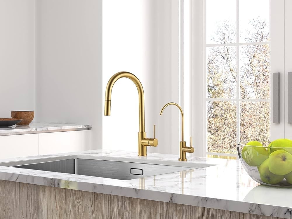 Modern kitchen sink with gold faucets, white countertop, and green apples in bowl by bright window with autumn trees visible outside