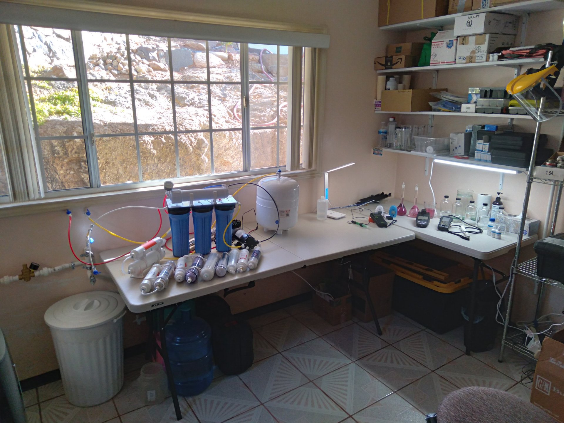 Home water filtration system laboratory setup with blue filter units on counter, shelving, and window overlooking outdoor trees