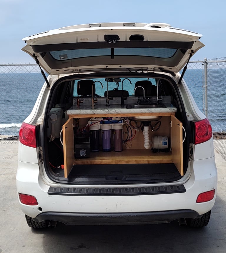 White minivan with open cargo area showing wooden storage shelves and camping supplies, parked at a coastal location