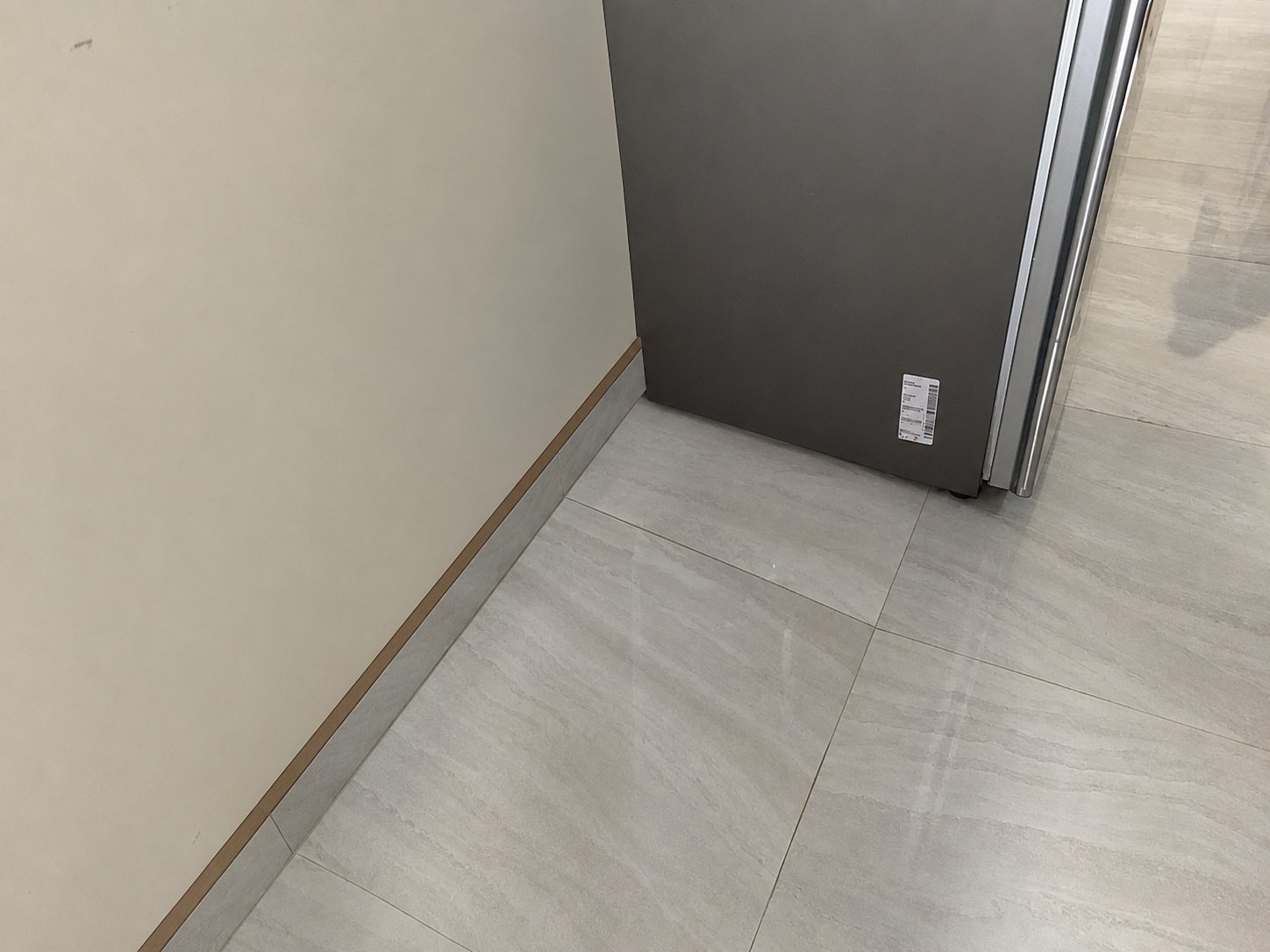 Corner of a room with light gray tile flooring, beige wall, baseboard trim, and a dark appliance partially visible with chrome trim and a label sticker