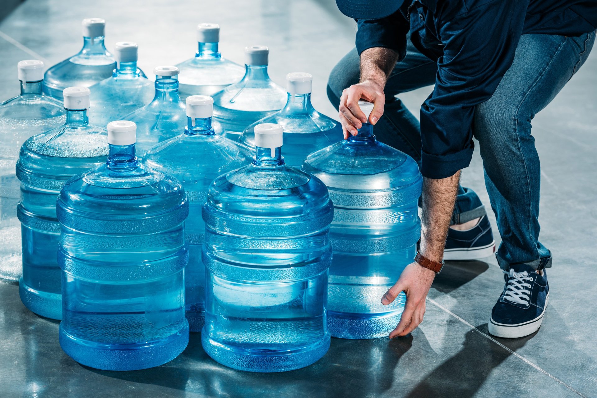 Person arranging multiple large blue water bottles and smaller water containers on a gray floor