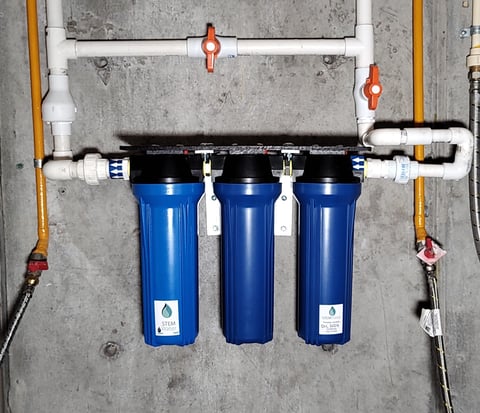 Three blue water filter canisters mounted horizontally under concrete with white and black PVC piping and red shut-off valves